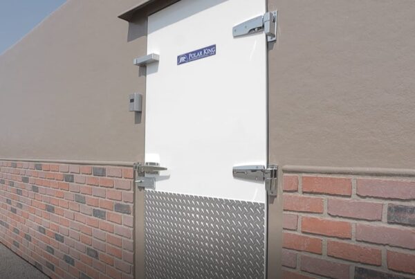 Walk in cooler with brown siding, white door, and brick overlayed with brown paint